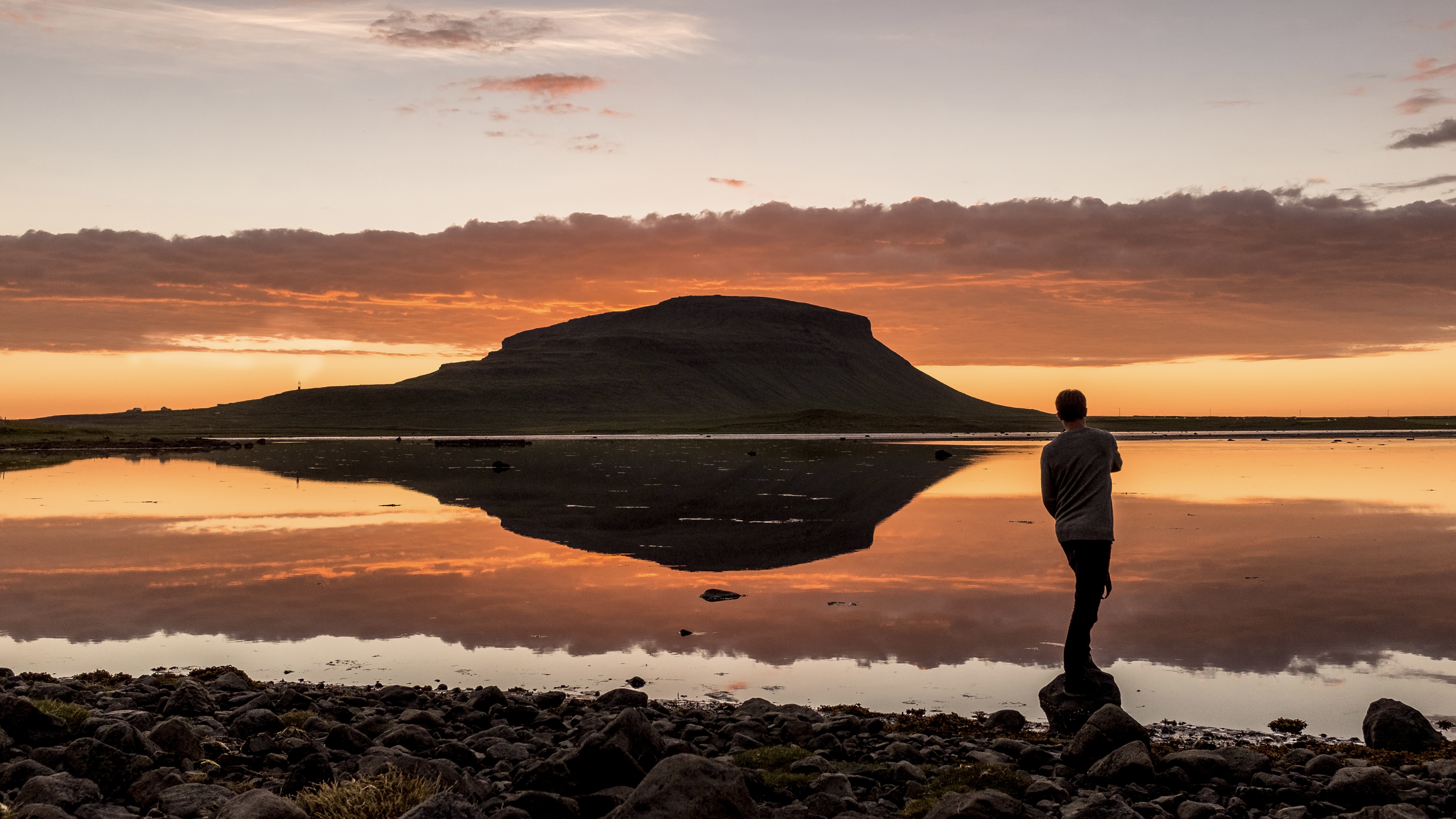 man_standing_on_a_rocky_shore_and_reflection_of_a_rock_peak_on_calm_water_at_sunset-4812x3208