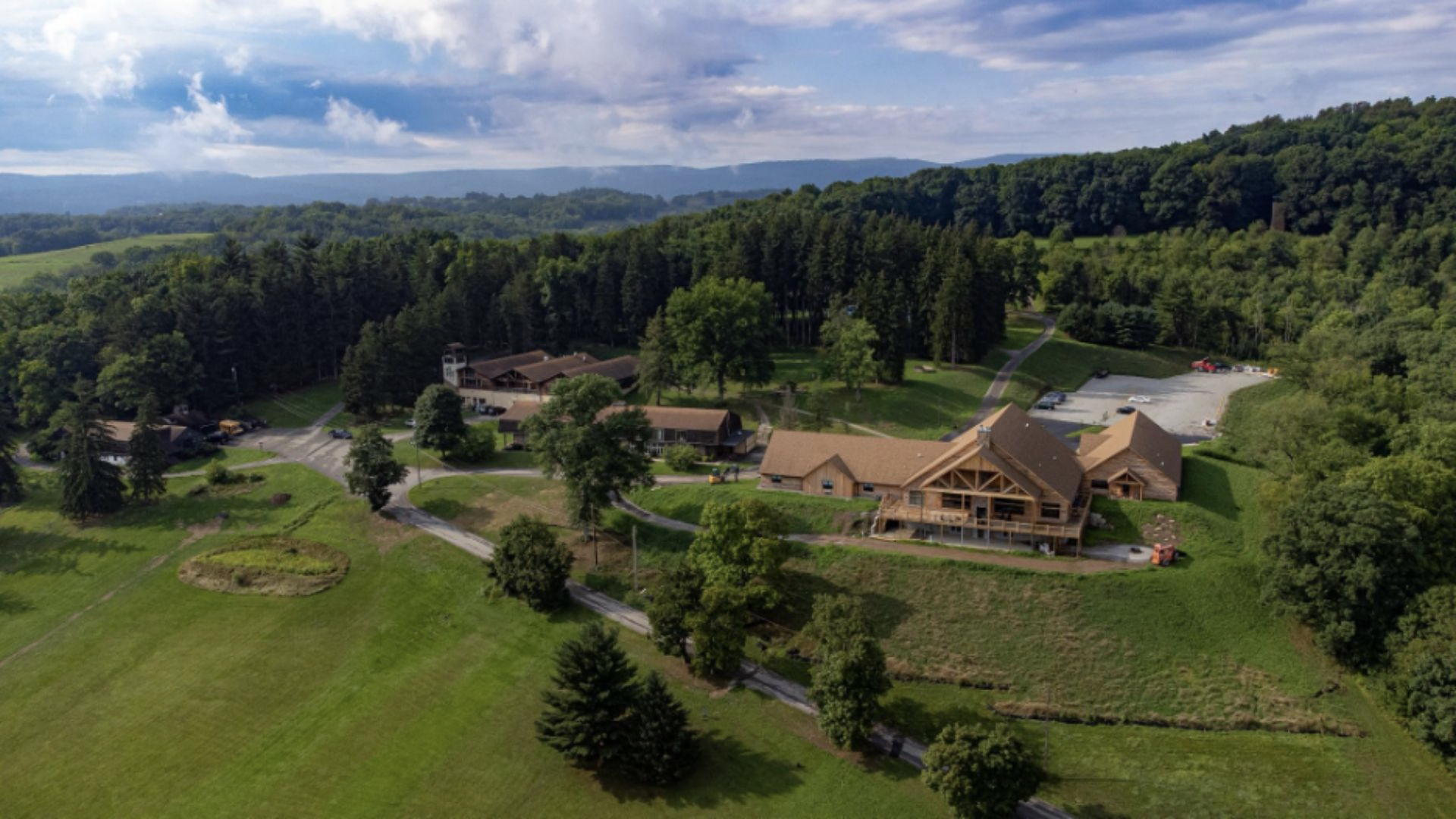 Aerial view of Ligonier Camp and Conference center, a rustic lodge nestled among lush trees and rolling hills. The scene conveys tranquility, with a clear blue sky above and expansive greenery.