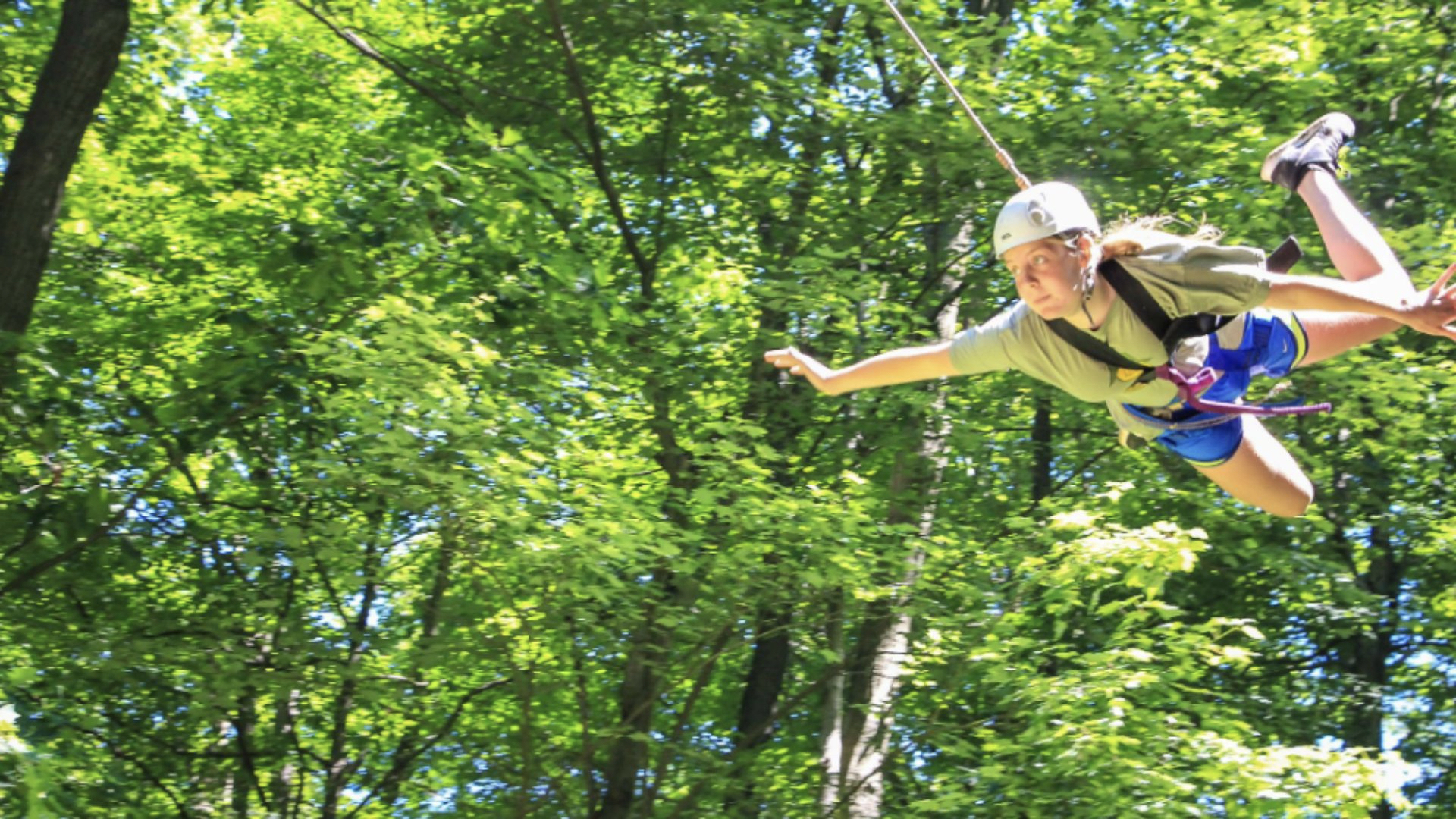 A person in a helmet and harness joyfully zip-lining through lush green trees on a sunny day.