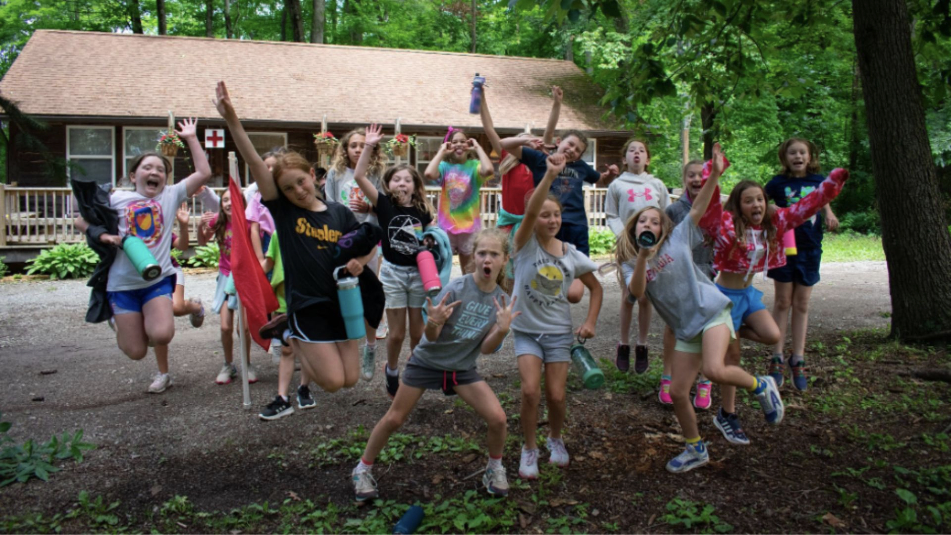 A group of excited children jumps enthusiastically in front of a rustic cabin.