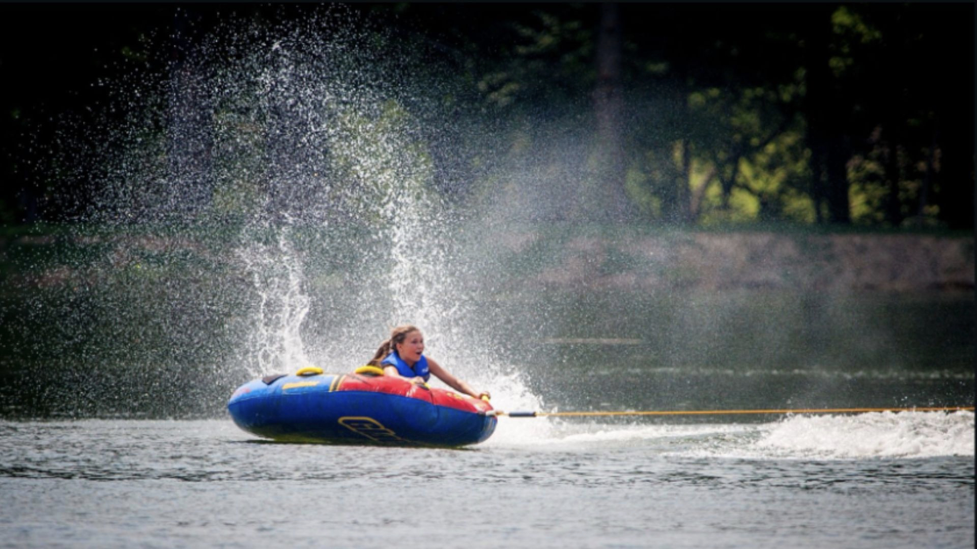 A person in a life vest rides a blue and yellow inflatable tube on a lake, creating a large spray.