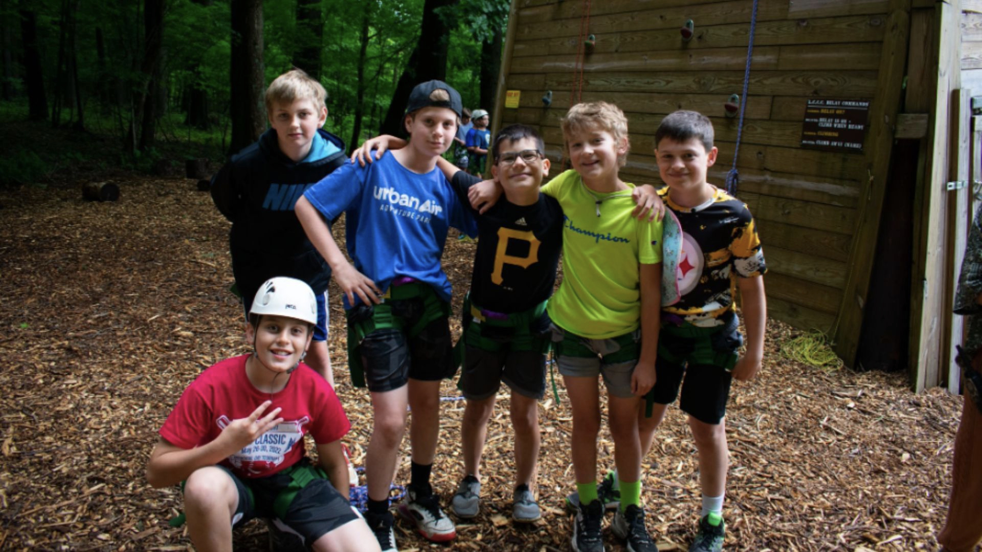 Six boys, smiling and wearing casual clothes, pose together in front of a wooden climbing wall. 