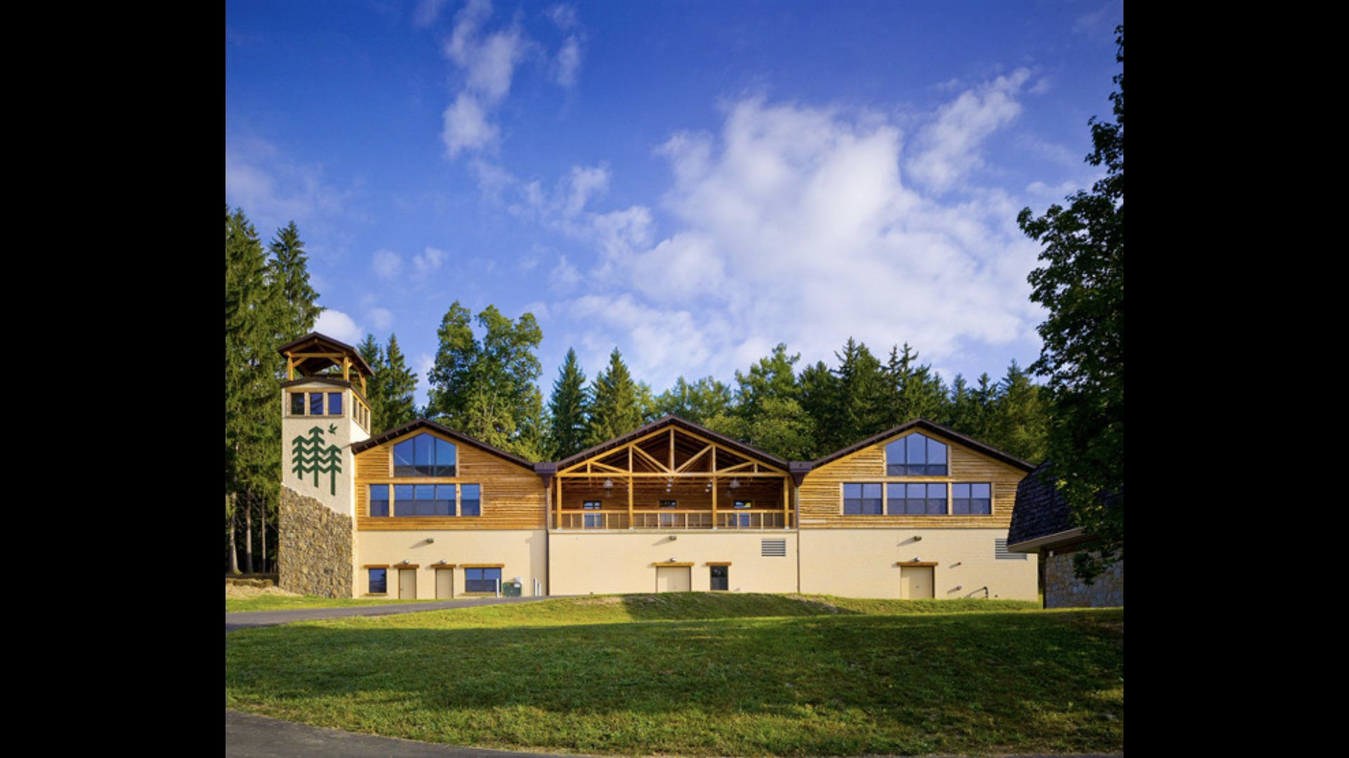 Ligonier Camp and Conference Center from the ground level. It is a large lodge with wooden accents and sits in front of a forest under a blue sky with clouds.