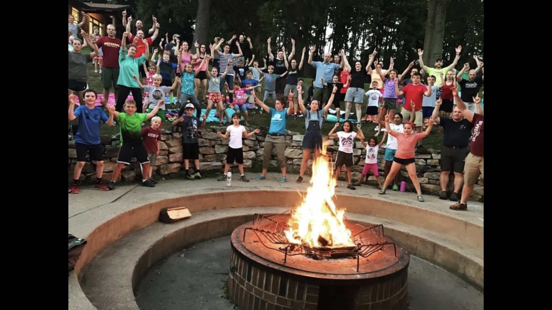 A large group of children and adults stand enthusiastically around a campfire, raising their arms in celebration.